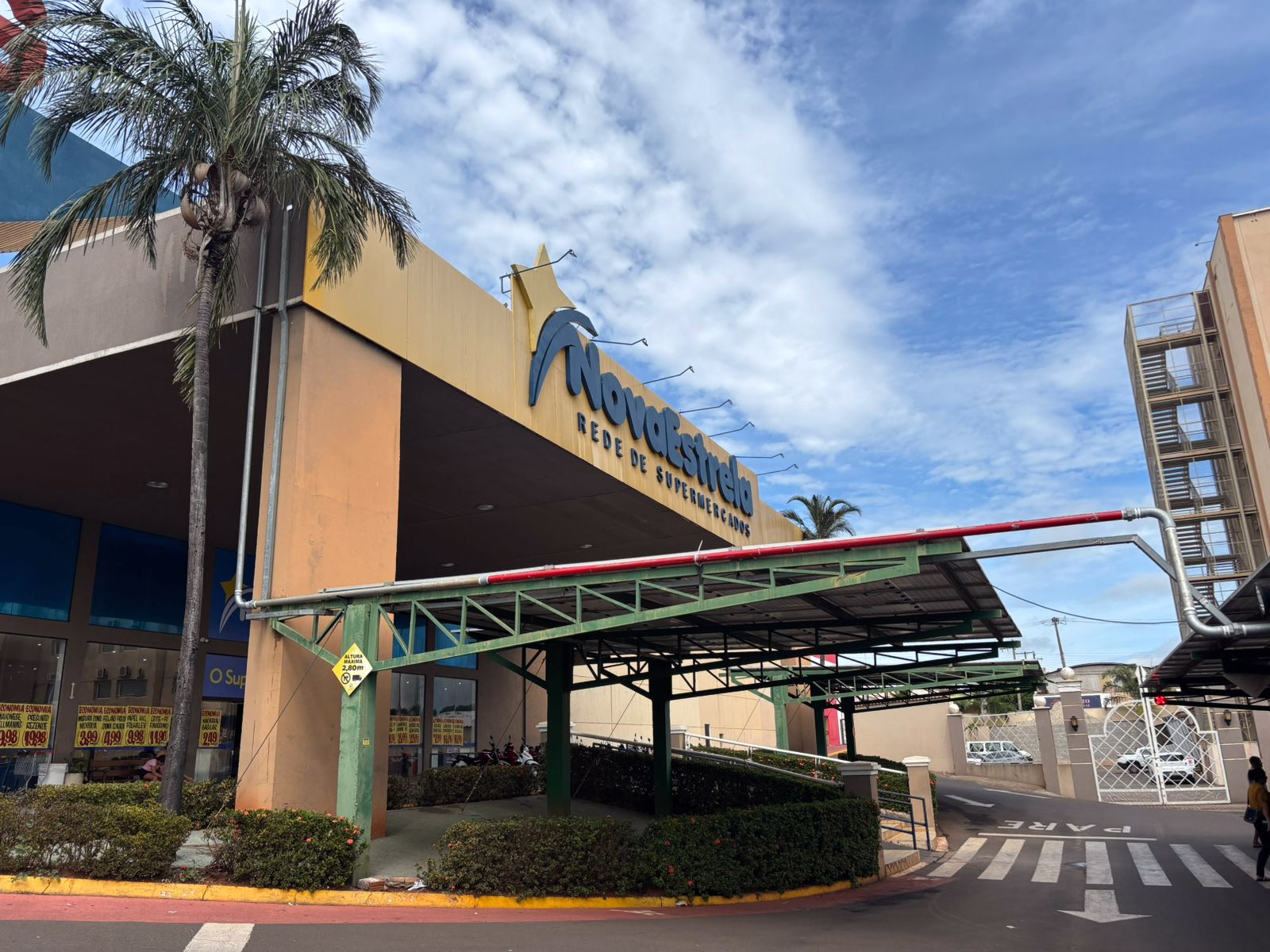 Entrada externa do supermercado Novostrela com toldo metálico verde e céu parcialmente nublado.