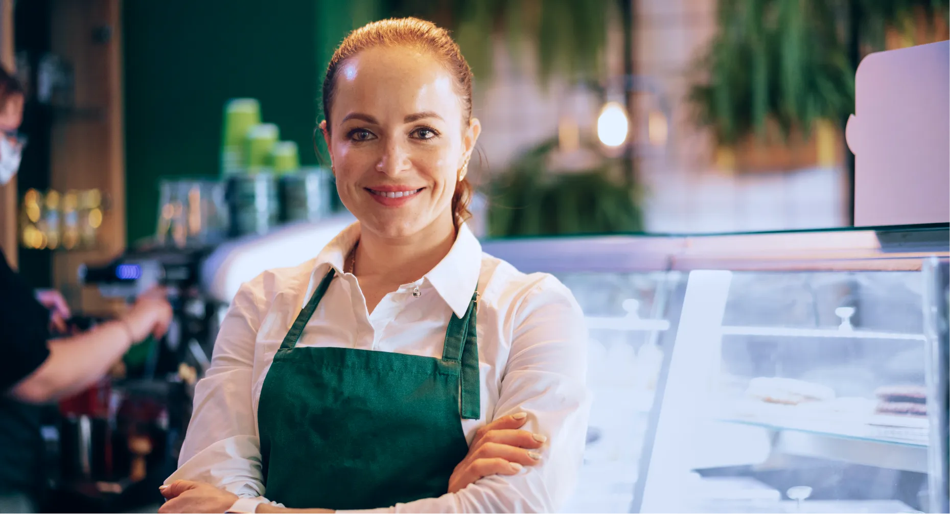 Mulher sorridente usando avental verde em uma cafeteria com vitrine de doces ao fundo.