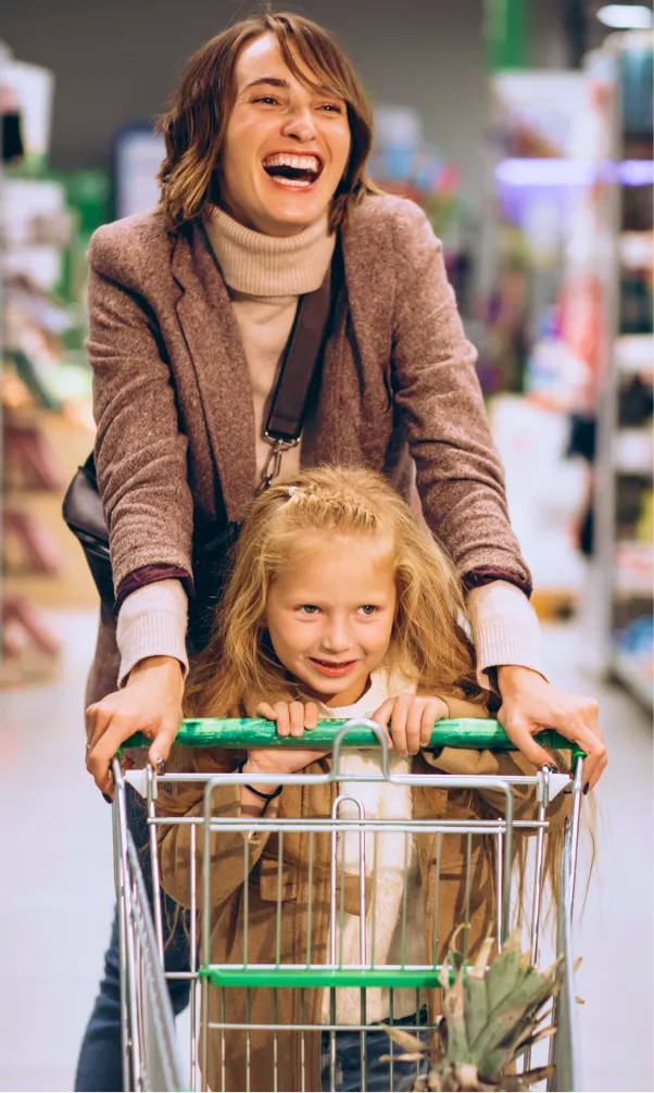 Mulher sorridente empurrando carrinho de compras com menina loira segurando o guidão em supermercado.