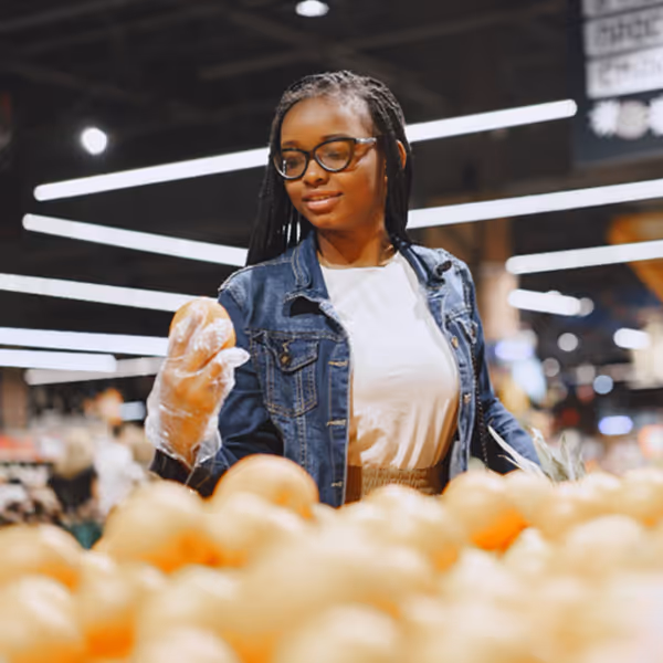 Mulher negra usando óculos, jaqueta jeans e luva plástica escolhendo cebolas em um supermercado.