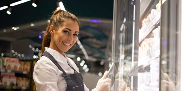 Mulher sorridente com avental e luvas abre porta de freezer em supermercado.