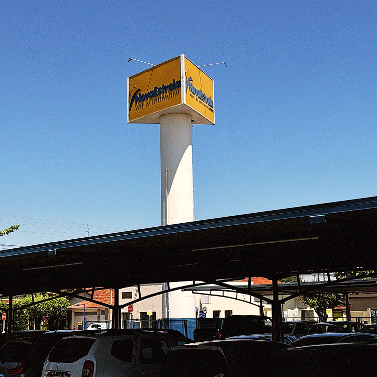 Estacionamento com carros sob cobertura e torre alta com placa amarela da loja NoveEstrelas contra céu azul.
